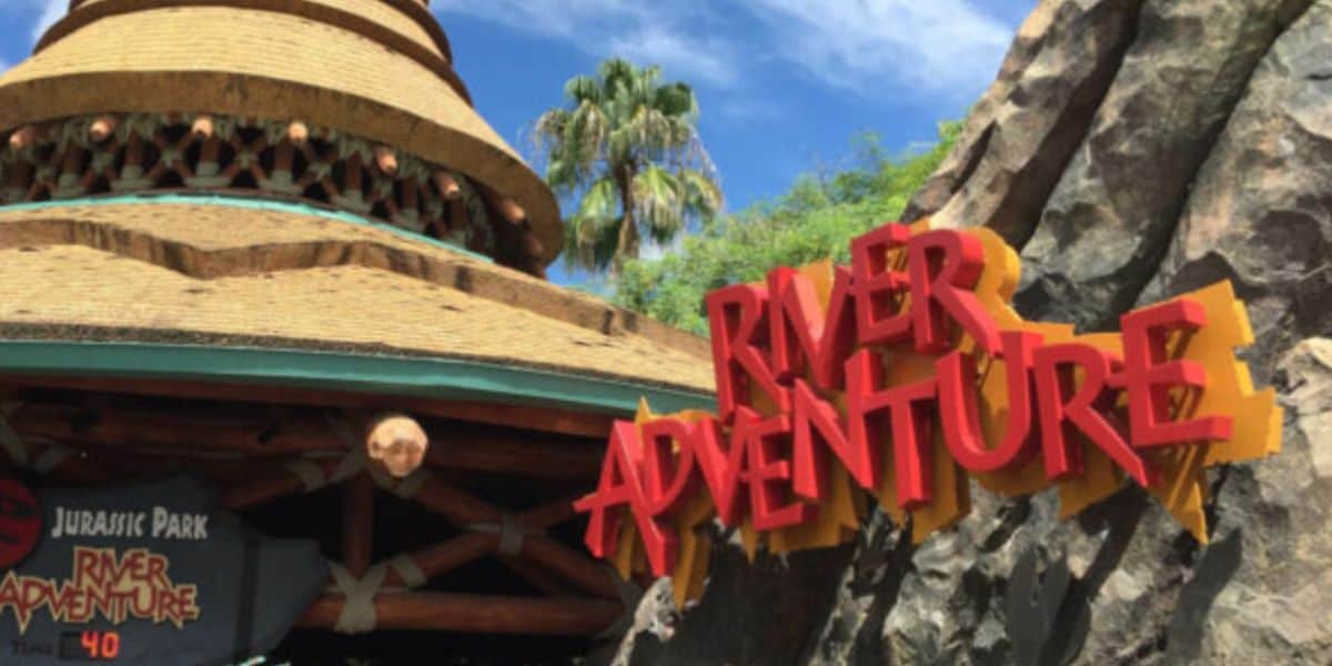The red and yellow River Adventure sign on a rock wall beside the thatched entrance building of Jurassic Park River Adventure, with a wait-time board reading 40