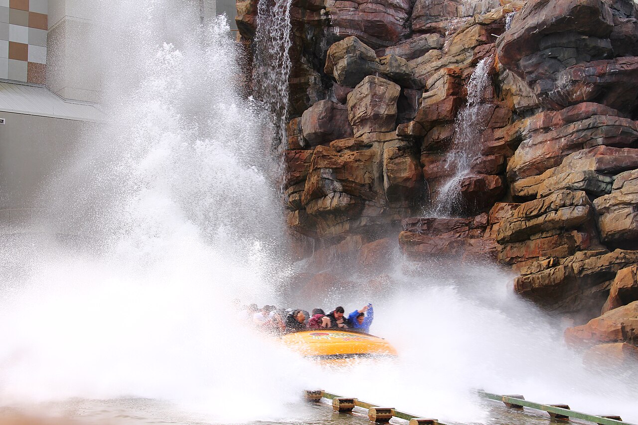 A yellow Jurassic Park River Adventure boat full of riders hits the water at the bottom of the drop, throwing up a huge wall of spray beside a rockwork cliff with waterfalls