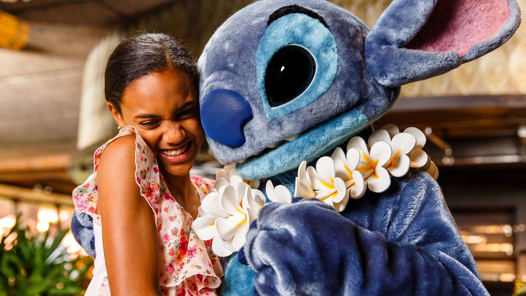 Stitch, wearing a white plumeria lei, hugs a laughing girl in a floral top inside 'Ohana