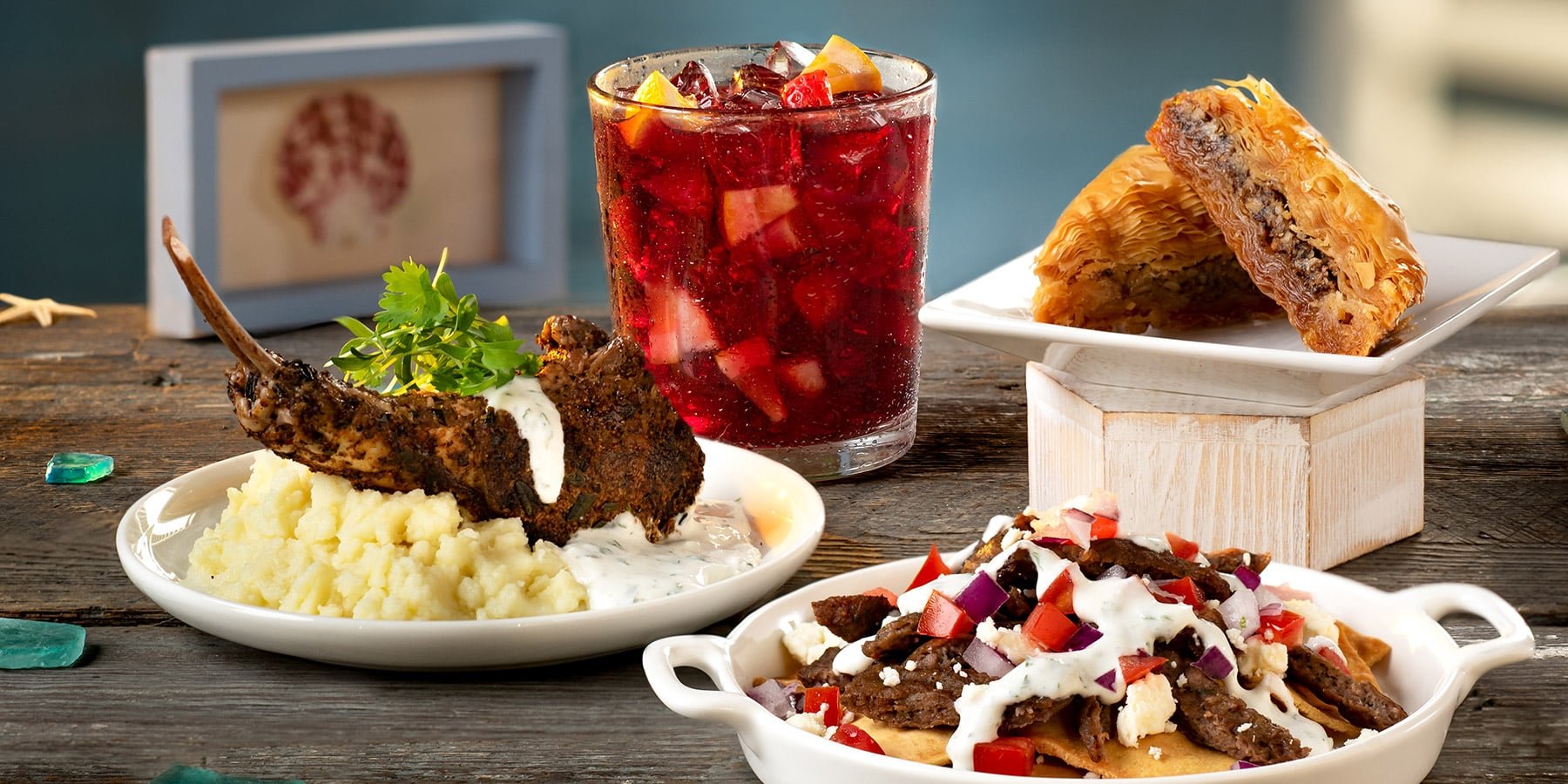Seven Seas Food Festival dishes on a wooden table: a lamb chop with a white yogurt sauce over mashed potatoes, an iced red drink with chopped fruit, two triangles of baklava and a dish of gyro nachos with feta, diced tomato and red onion