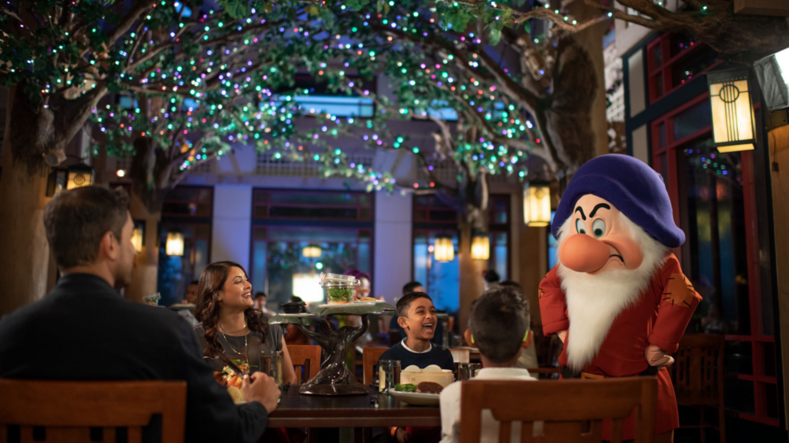 Grumpy the dwarf, hands on hips, scowls at a laughing family at a table beneath a canopy of twinkling lit trees at Story Book Dining at Artist Point
