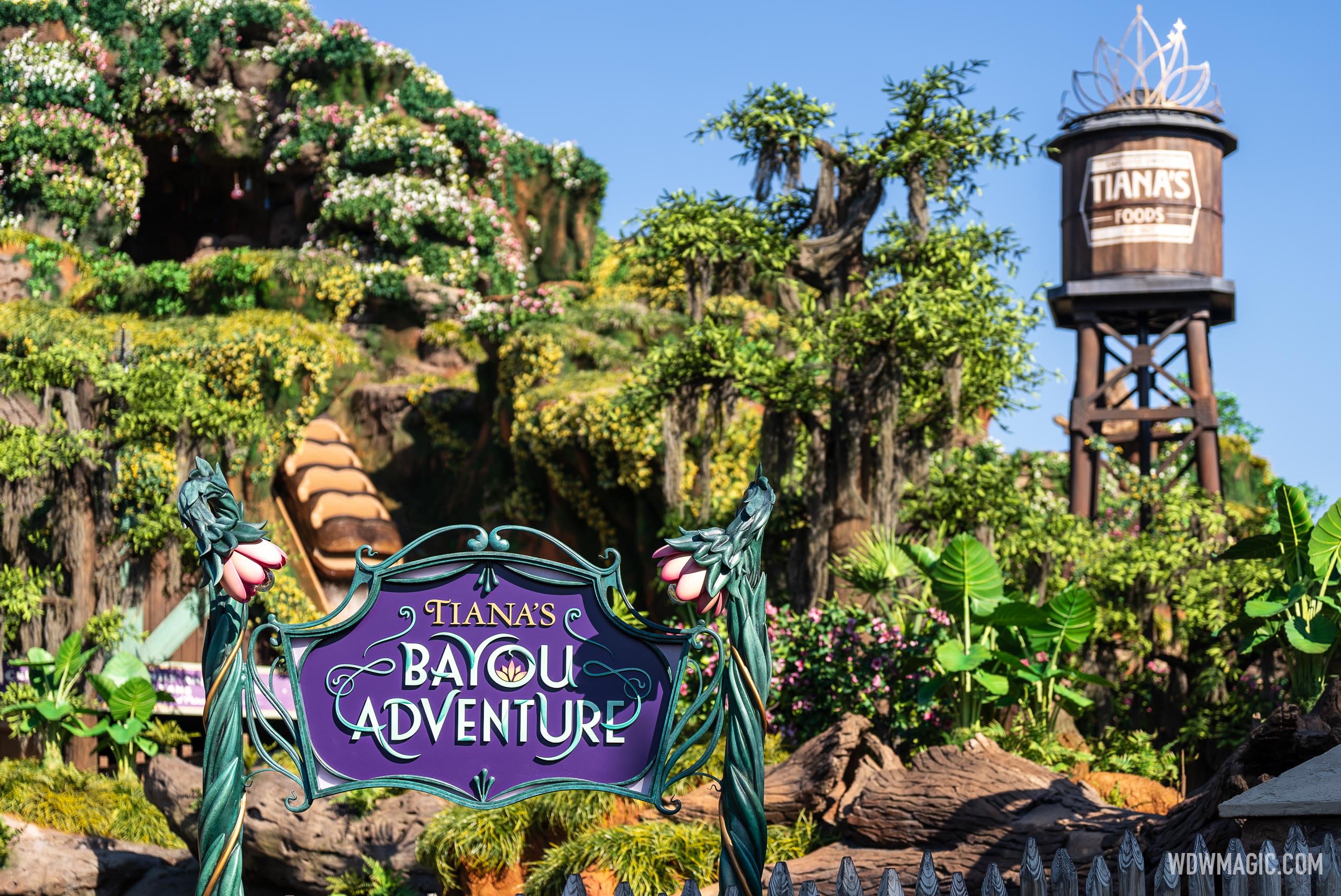 The purple Tiana's Bayou Adventure sign in front of the flower-covered mountain, with a log on the drop and the Tiana's Foods water tower at right