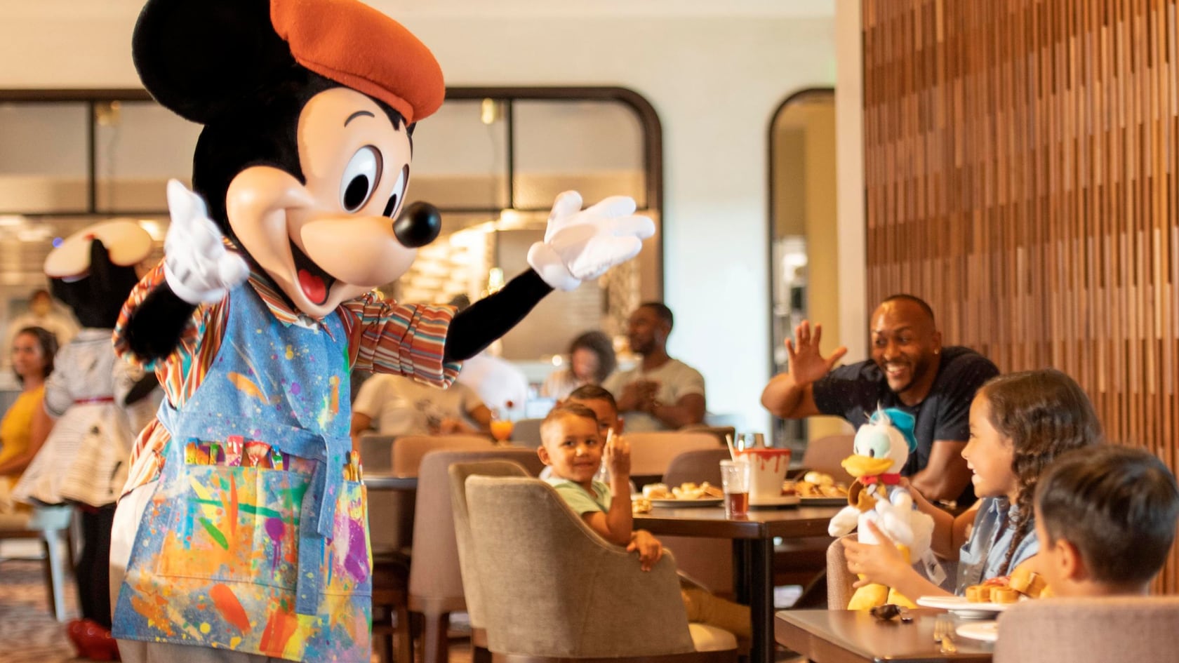Mickey Mouse in an orange beret and a paint-splattered artist's smock waves to cheering children and a man at a table at Topolino's Terrace, with Minnie Mouse in the background at left