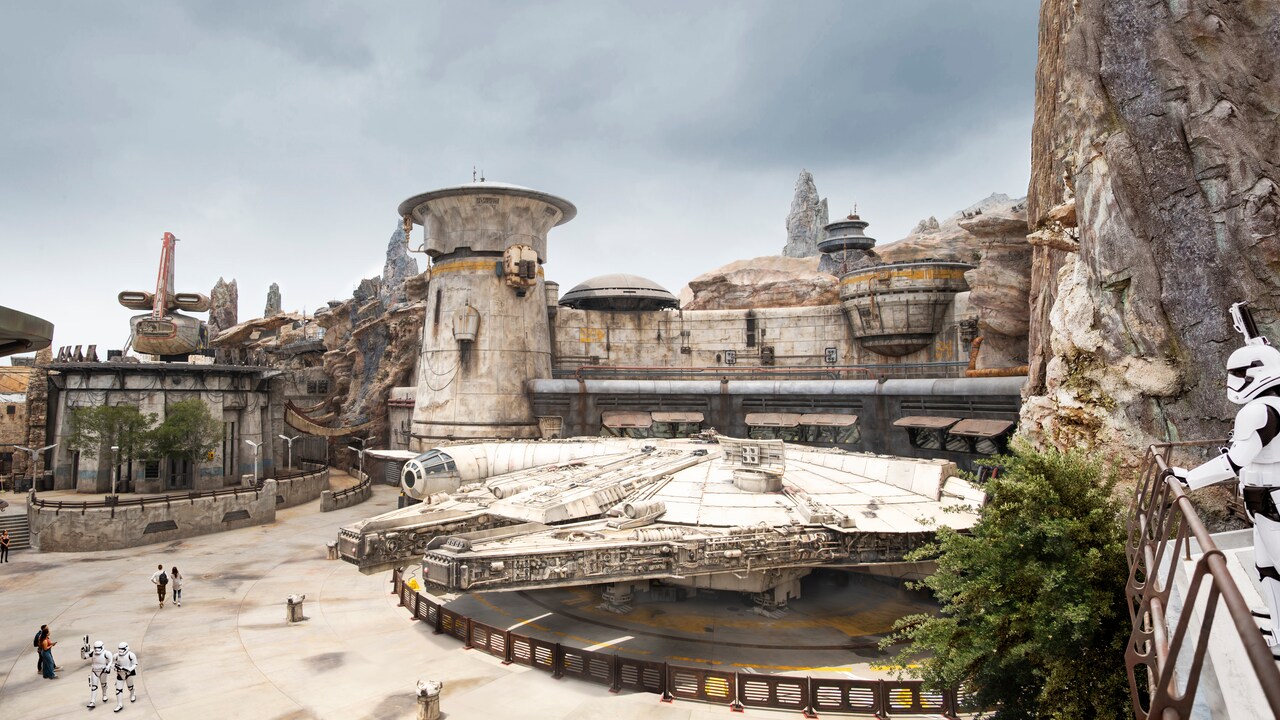 The Millennium Falcon parked in front of the rock spires and domed buildings of Black Spire Outpost, with two stormtroopers walking past guests in the courtyard