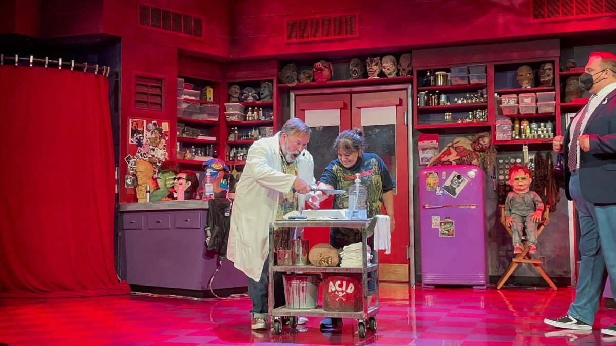 A host in a lab coat and an audience volunteer at a steel cart on a red-lit stage lined with shelves of monster masks and props, with a second host standing at right