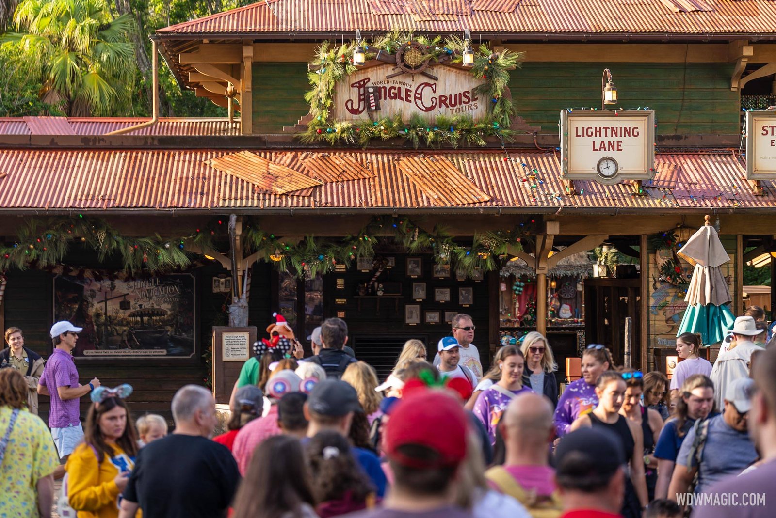 Guests walk past the Jingle Cruise entrance building, with a garland-framed World-Famous Jingle Cruise Tours sign on the roofline and a Lightning Lane sign hanging to the right