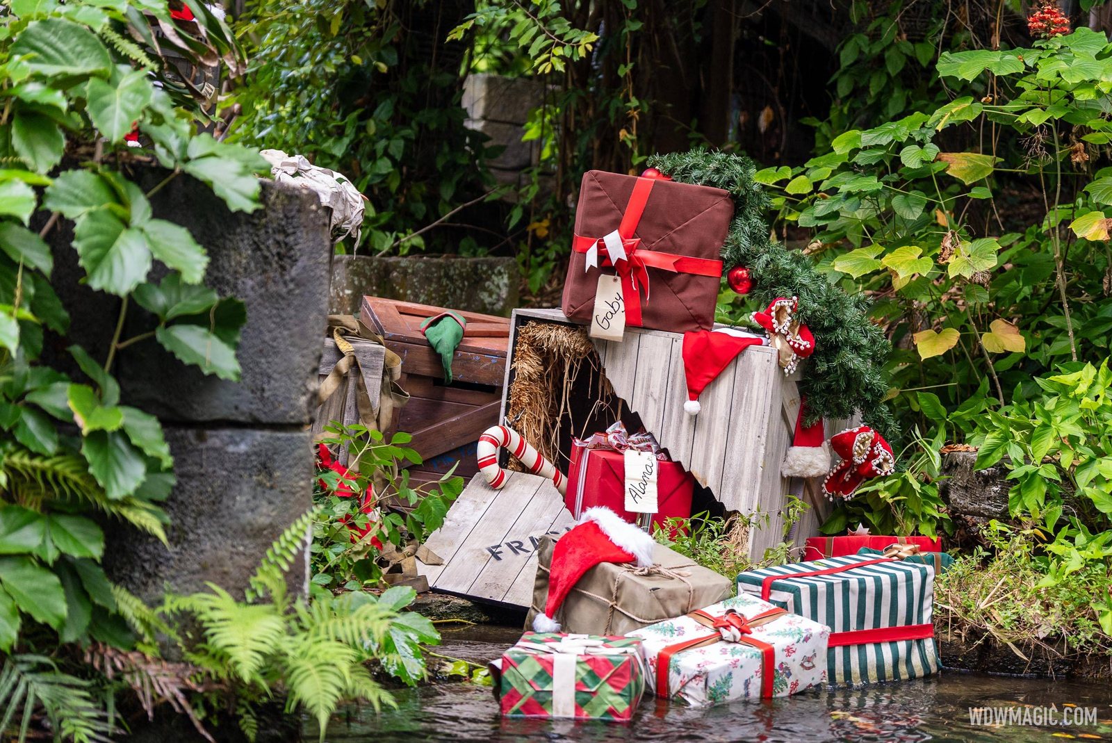 A pile of wrapped presents in red, white and green paper, along with stockings and a Santa hat, stacked on the riverbank beside the Jungle Cruise water