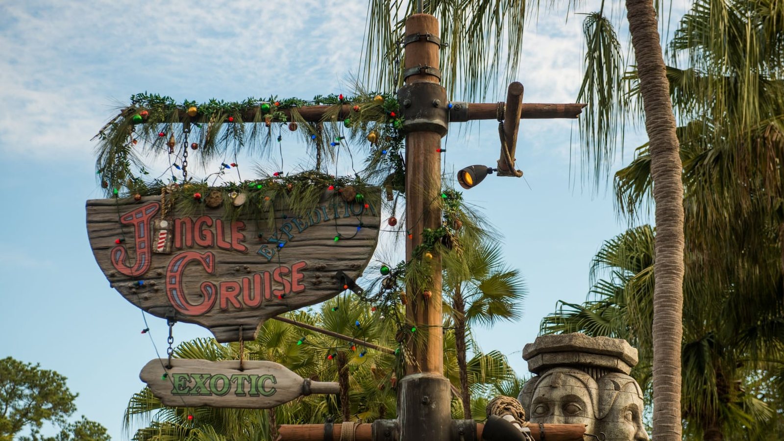 The Jingle Cruise sign wrapped in garland and colored lights, hanging from a wooden post with an Exotic arrow sign below, palm trees and blue sky behind
