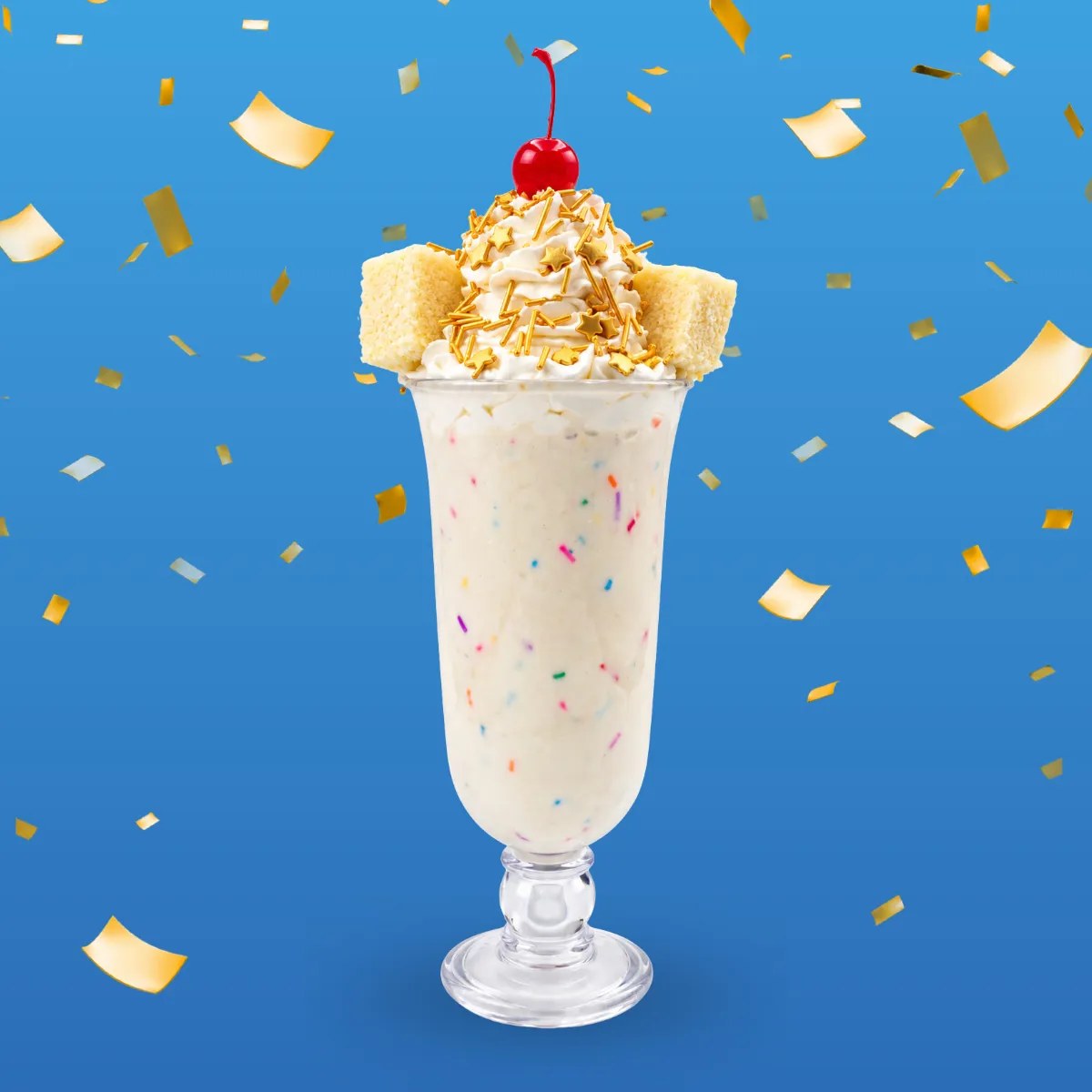 A tall footed glass of vanilla milkshake speckled with rainbow sprinkles, topped with whipped cream, gold star sprinkles, two vanilla cake cubes and a red cherry, against a blue background with gold confetti