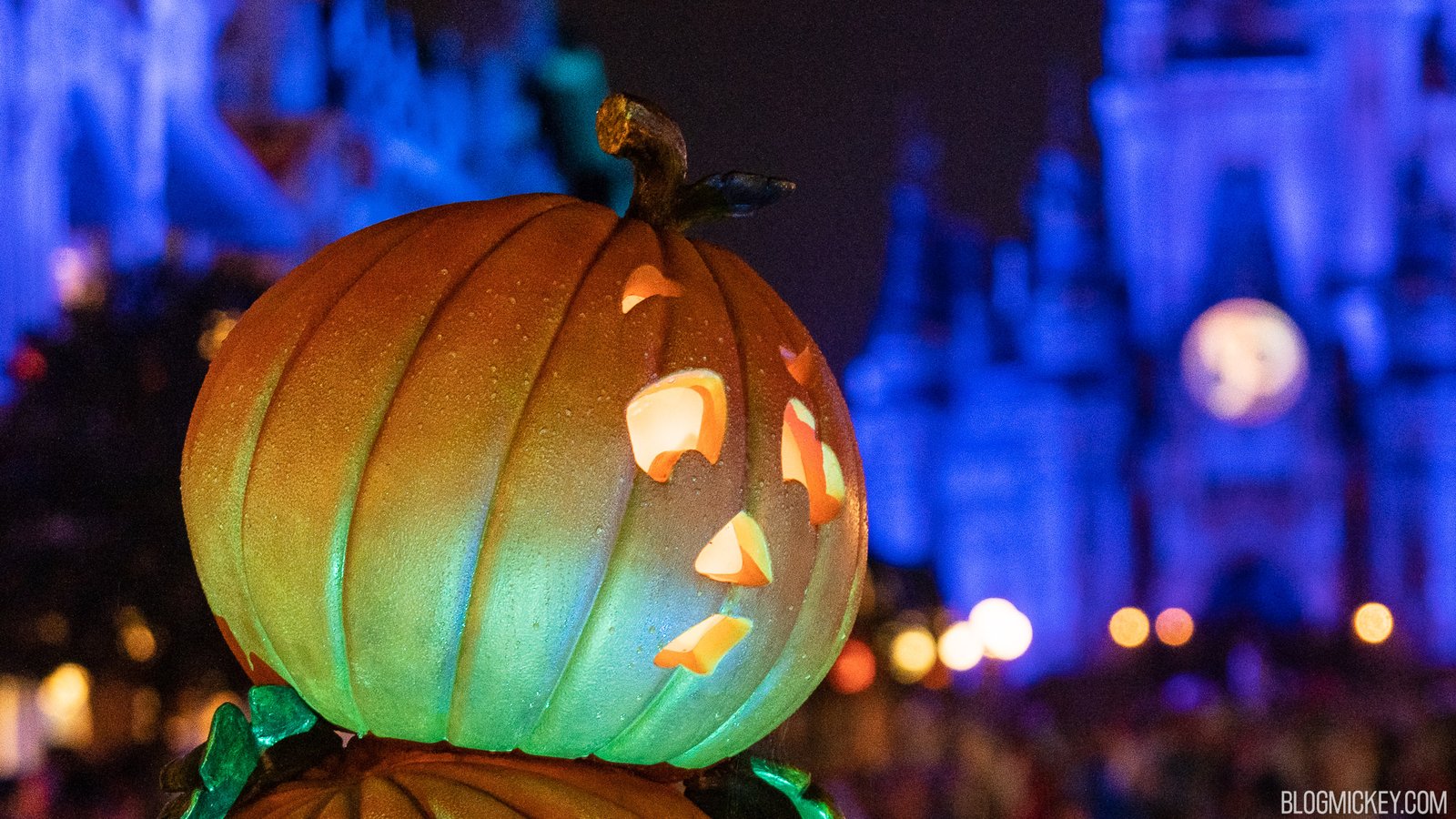 A rain-beaded jack-o'-lantern lit green and orange in front of a blurred, blue-lit Cinderella Castle at night
