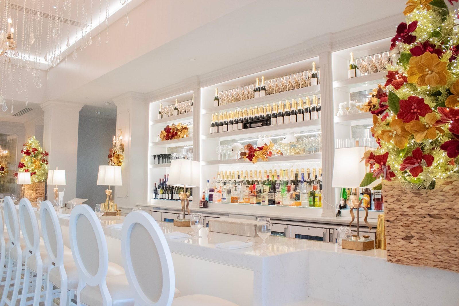 A white bar with round-backed white barstools, backlit shelves of champagne bottles and glassware, crystal chandeliers and lighted holiday trees covered in flowers