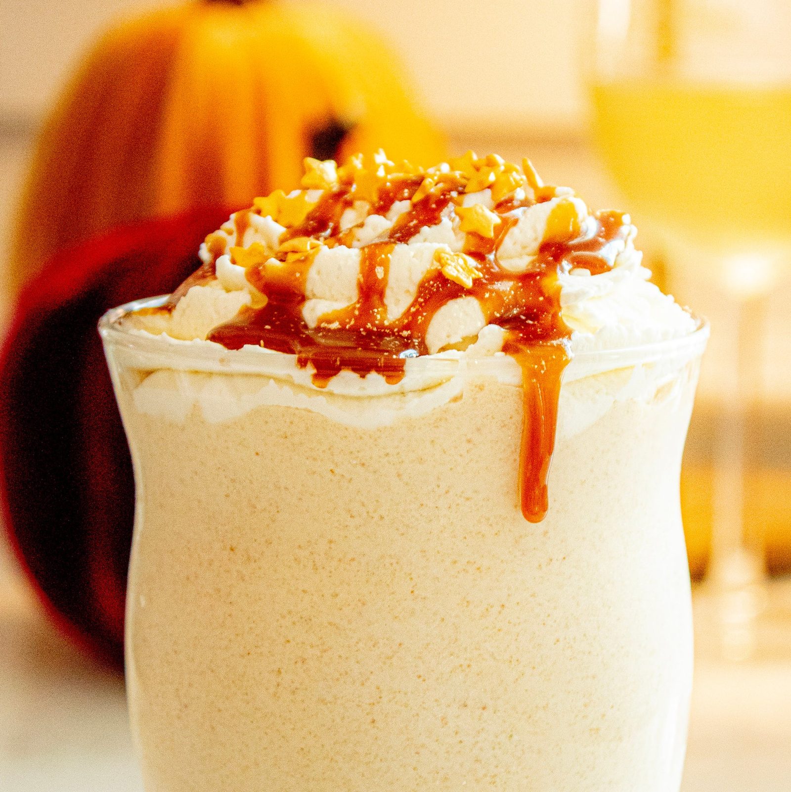 A close-up of a tall milkshake topped with whipped cream, caramel drizzle and gold star sprinkles, with a pumpkin blurred behind it
