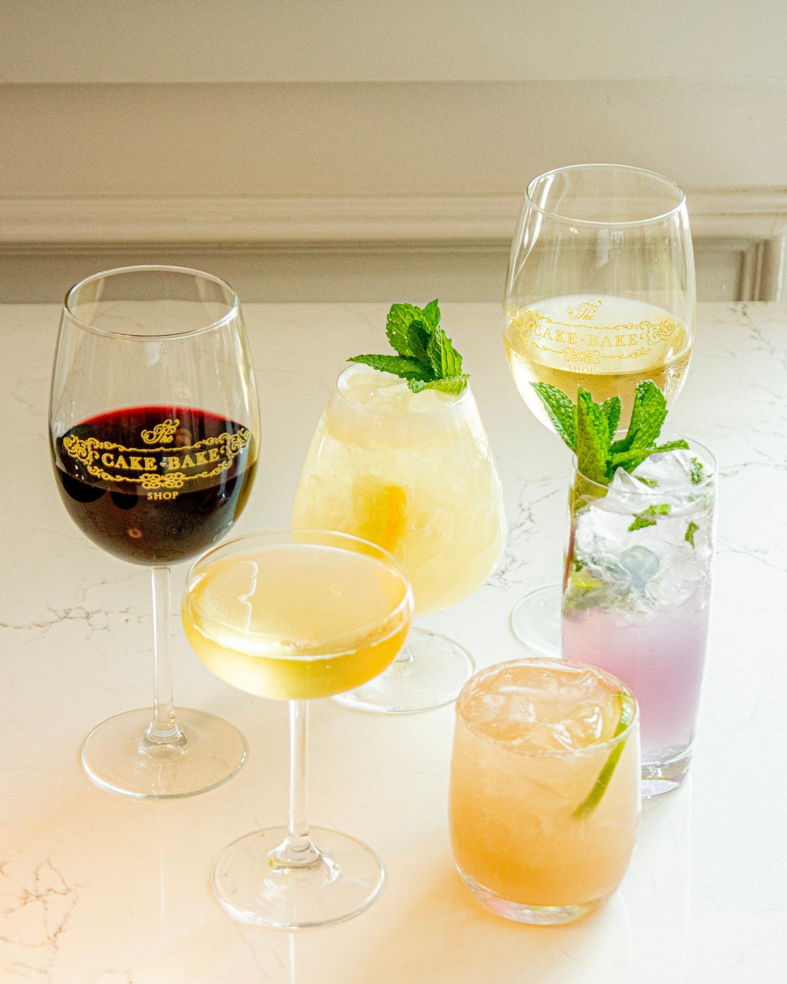 A glass of red wine and a glass of white wine printed with the gold Cake Bake Shop logo, a coupe, a mint-topped cocktail in a stemmed glass, a mint-topped lavender highball and a pink cocktail with lime on a marble counter