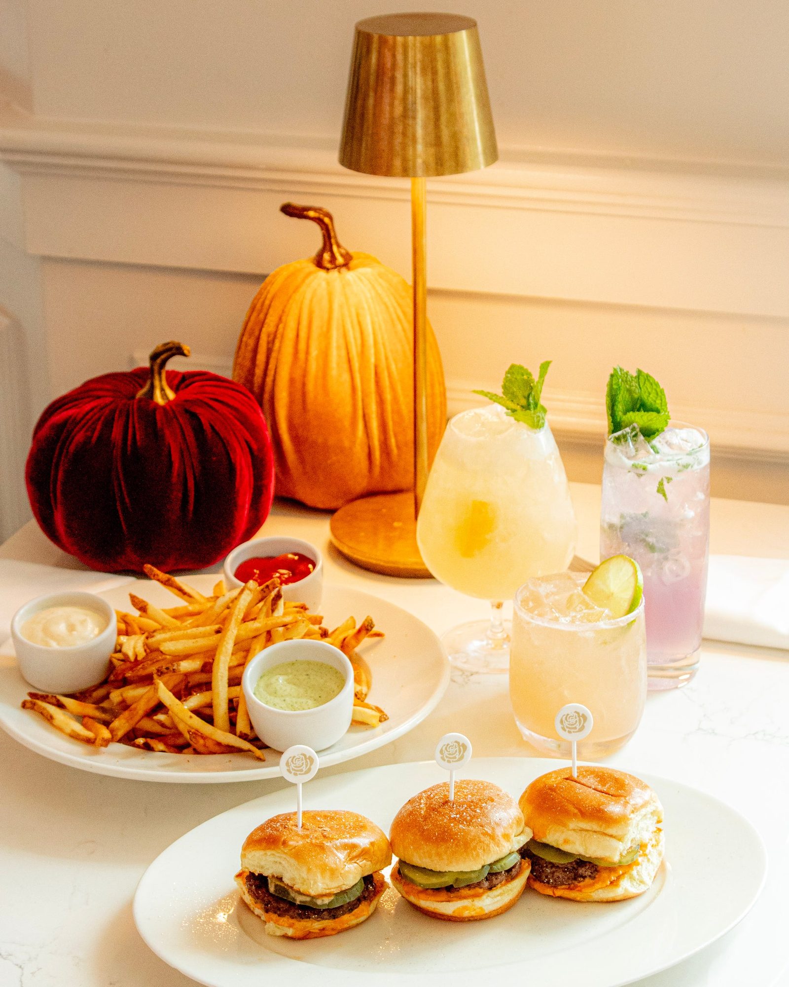 A plate of fries with three dipping sauces, a plate of three sliders with rose-topped picks, and three cocktails on a white marble table beside a gold lamp and velvet pumpkins