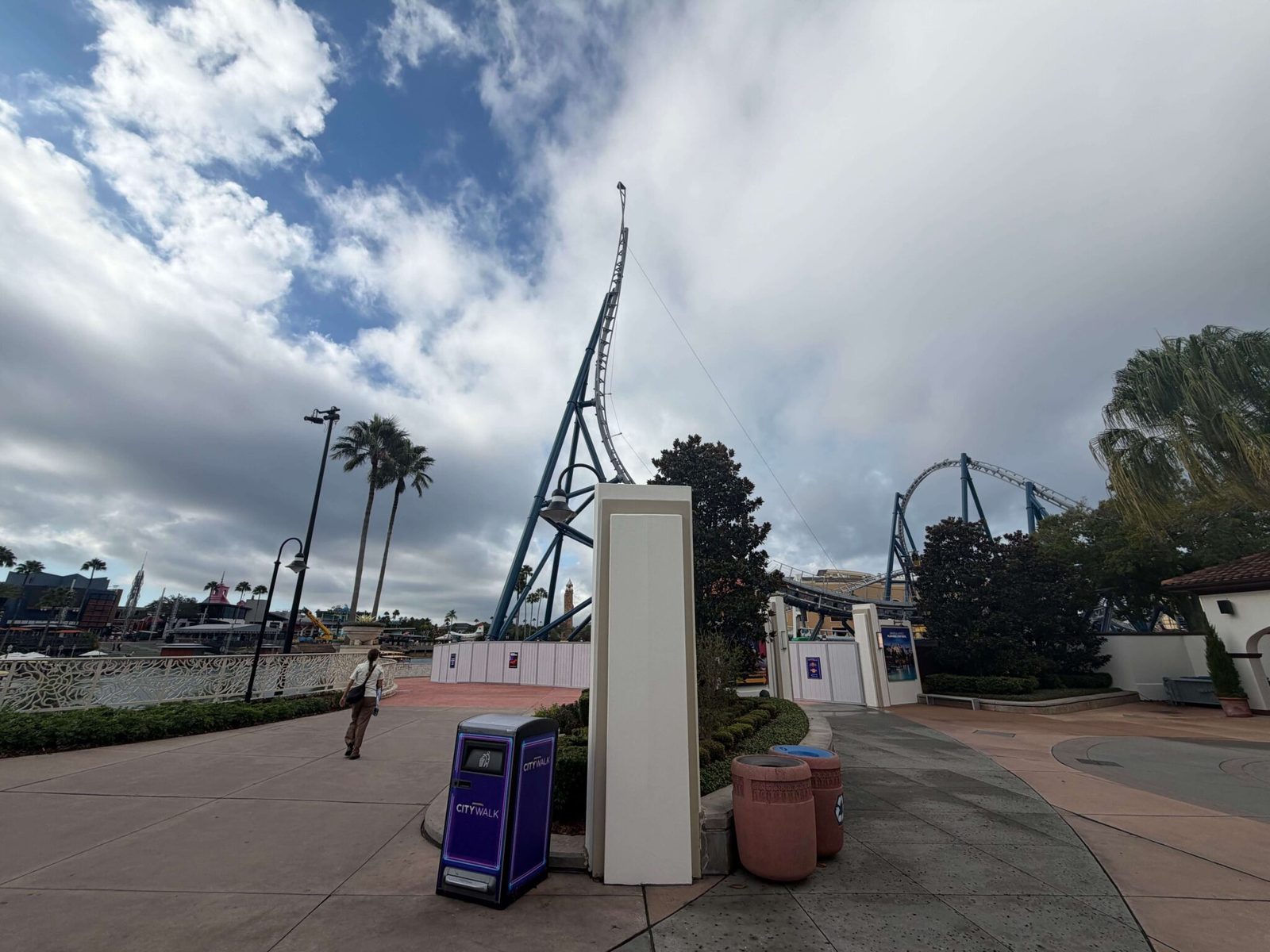 A wide CityWalk walkway beside the waterfront railing, with a CityWalk trash bin, two terracotta bins and a white pillar, and Hollywood Drift's tall vertical spike on blue supports rising behind construction walls under a cloudy sky