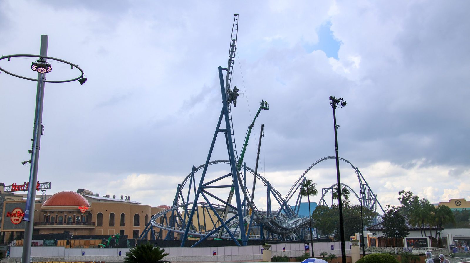 Wide view of the blue Hollywood Drift coaster structure under a cloudy sky, with a small dark vehicle high on the vertical spike, a boom lift beside it, and the Hard Rock Cafe dome at left