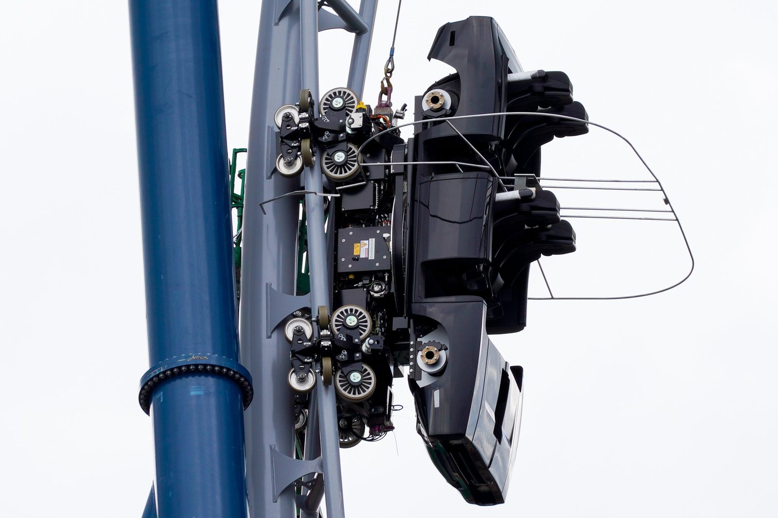 A black coaster test vehicle hanging on near-vertical gray track beside a blue support column, with metal clearance frames around it and a wire rope above