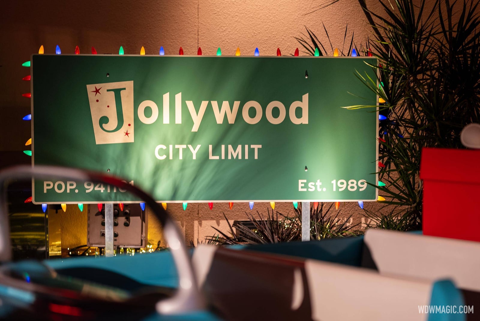A green road-style sign reading Jollywood City Limit and Est. 1989, outlined in colored string lights, with a blurred teal vintage car and gift boxes in the foreground