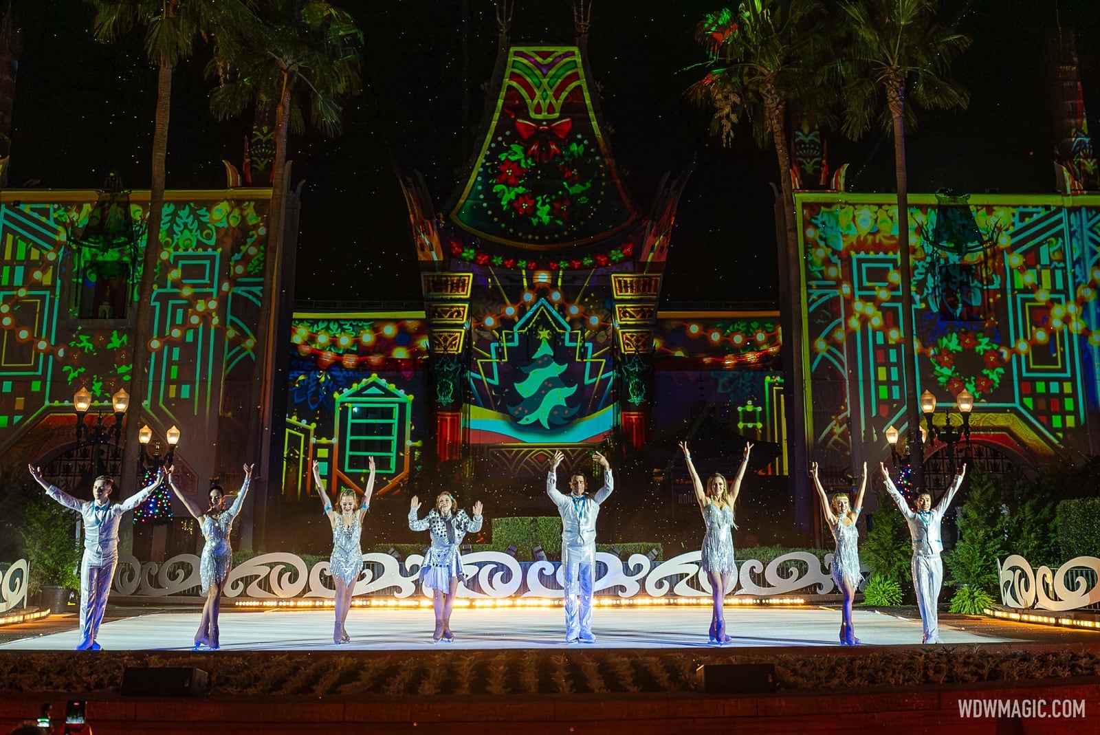 A line of skaters in white and silver costumes raise their arms on an outdoor rink in front of the Chinese Theater, which is lit with green and red holiday projections