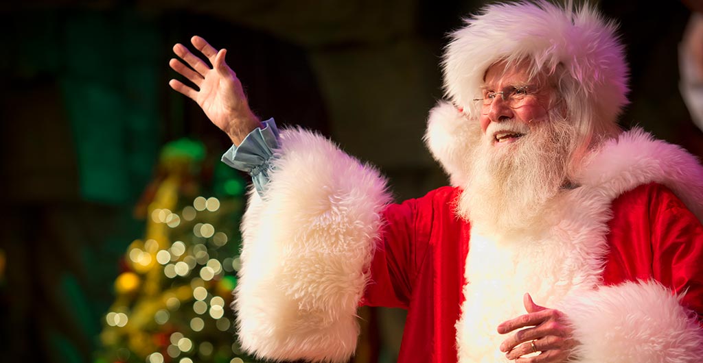 Santa Claus in a red suit with white fur trim waves with one raised hand in front of a lit Christmas tree