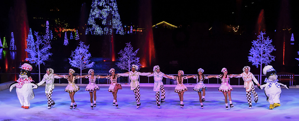 A line of ice skaters in white costumes with pink and black checks, joined by two penguin characters, skates across a lit rink in front of blue-lit trees and a large Christmas tree