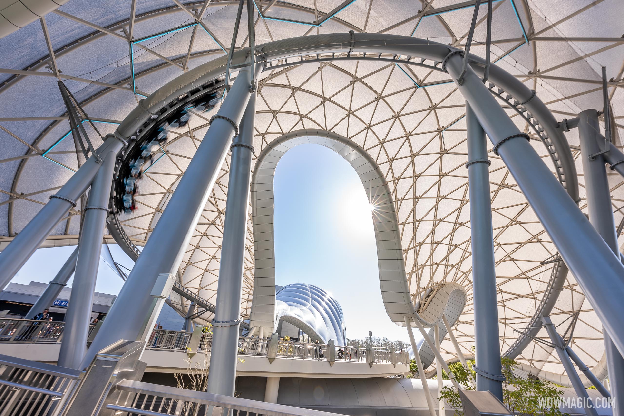A TRON Lightcycle / Run train streaks along the track under the ride's white lattice canopy at Magic Kingdom, with a bright sky visible through a looping arch