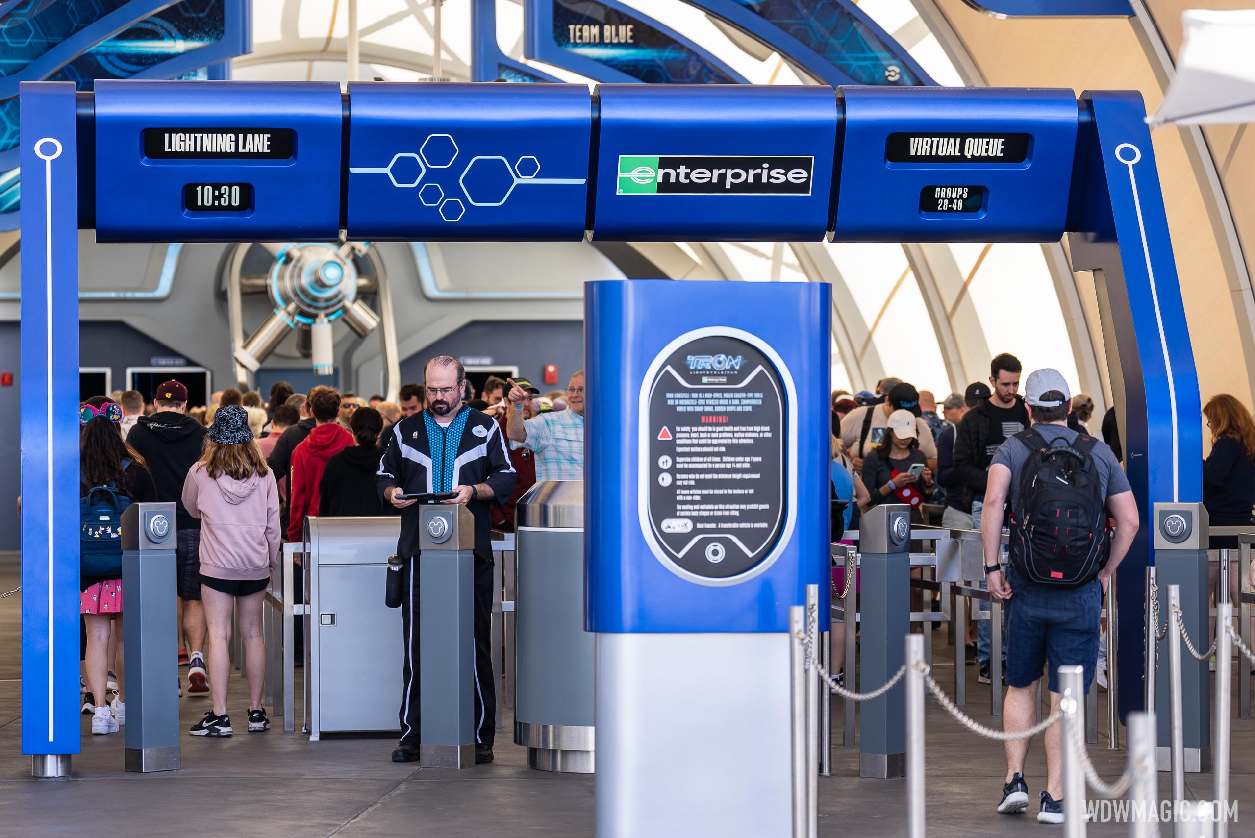 Guests pass through the TRON Lightcycle / Run entrance gates beneath a blue overhead sign reading Lightning Lane 10:30 on the left and Virtual Queue Groups 28-40 on the right, with a cast member holding a tablet and a ride information sign in front