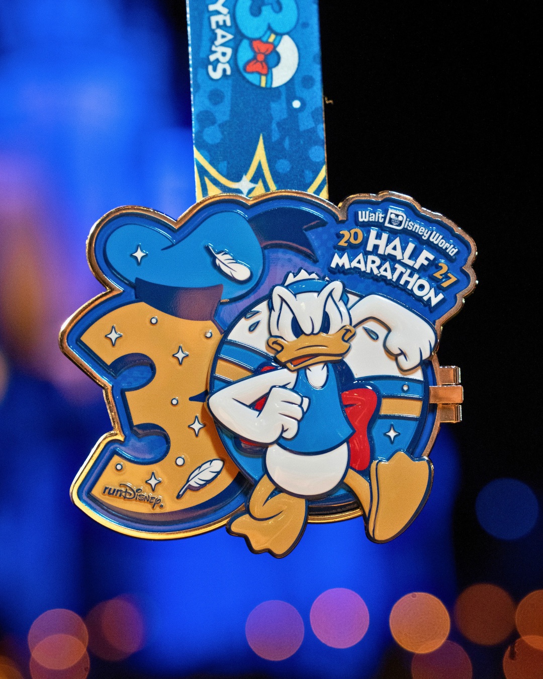 A blue and gold medal shaped like the number 30, with Donald Duck's blue sailor cap on top of the 3 and a scowling Donald running across the 0, which has a hinge on its right edge, hanging from a blue ribbon reading 30 Years