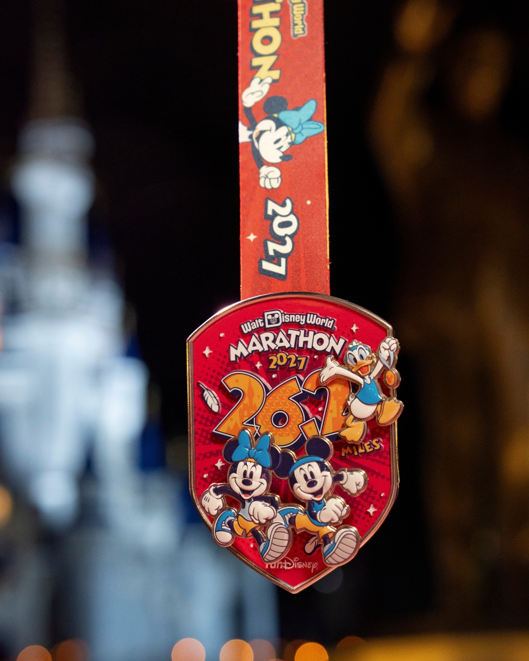 A red shield-shaped Walt Disney World Marathon 2027 medal showing Minnie and Mickey Mouse running in sneakers beneath large orange 26.2 numerals, with Donald Duck in the upper right holding up a medal, hanging from a red ribbon