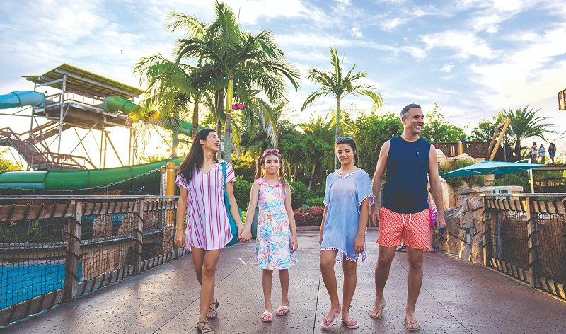 A smiling family of four in swimwear and cover-ups walk across a bridge at Aquatica, a mother holding her younger daughter's hand, with green water slides and palm trees behind them