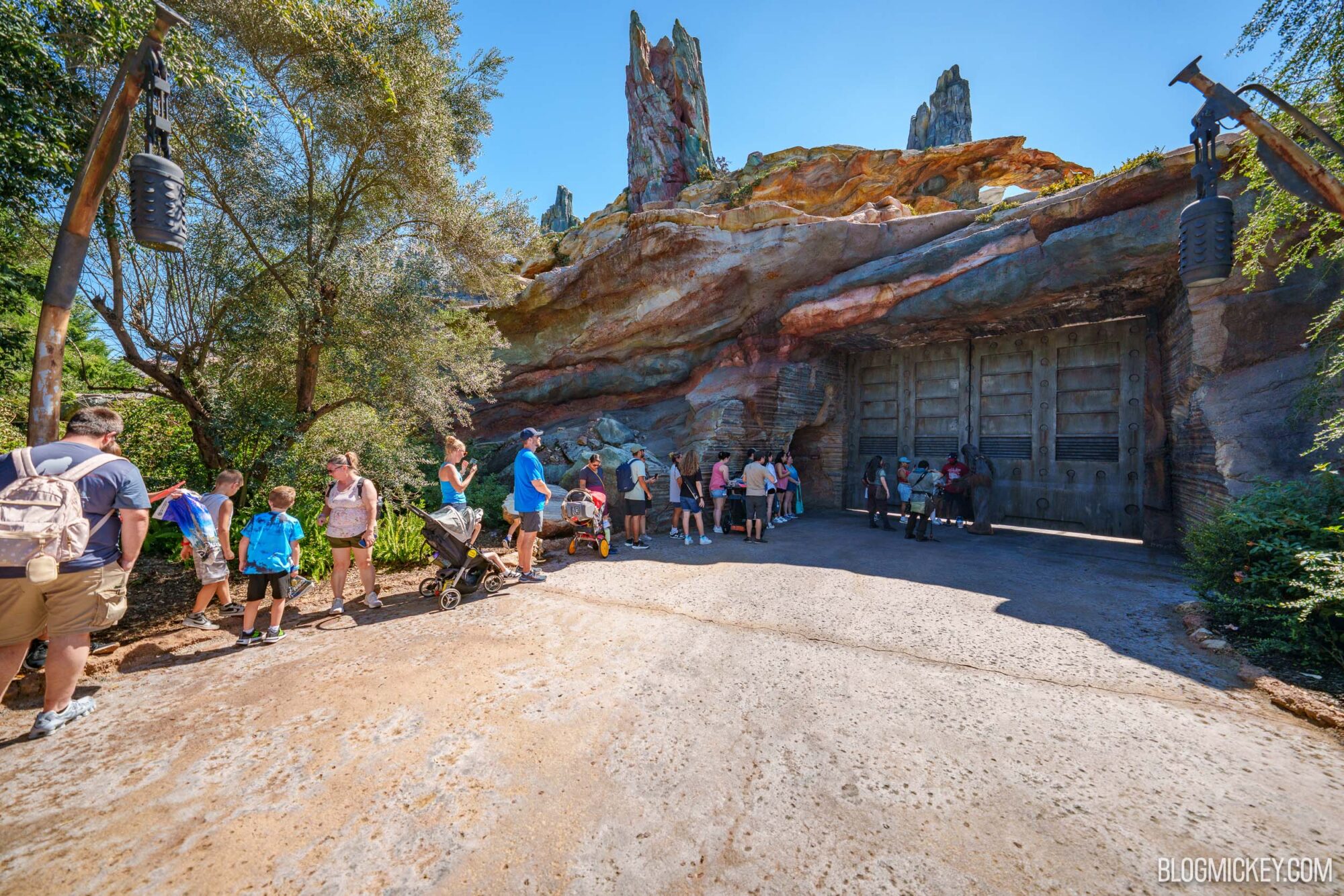 A line of guests with strollers waits along a path beside trees, leading to Chewbacca standing in front of a large metal gate set into colorful layered rockwork with tall rock spires above