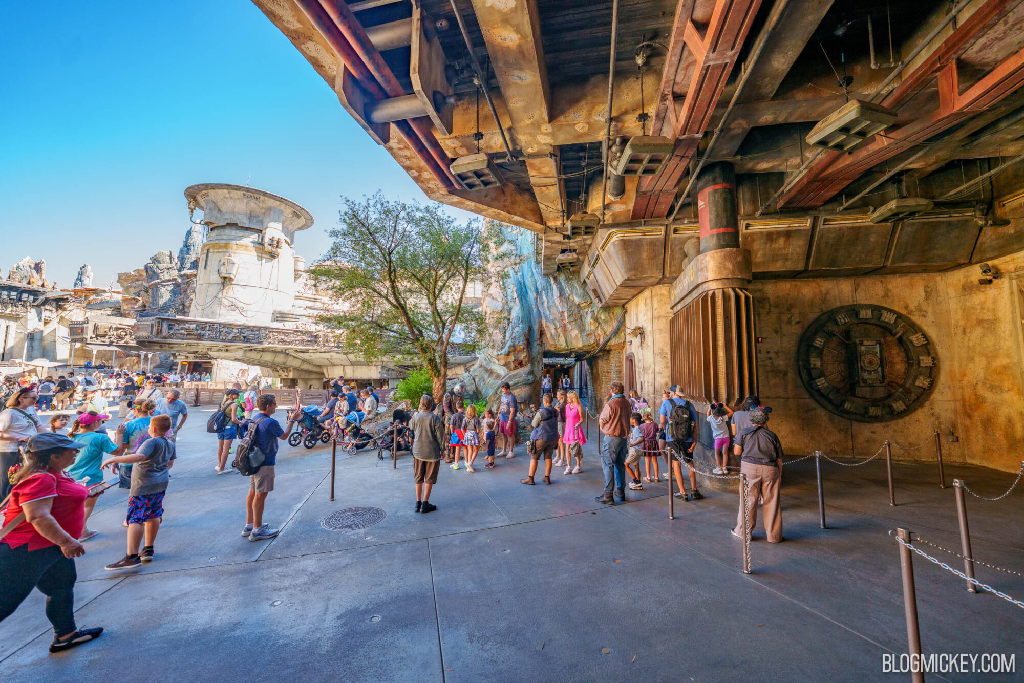Guests stand in a line marked by brown posts and chains under a huge rusted metal overhang, with a white tower and a tree in the background
