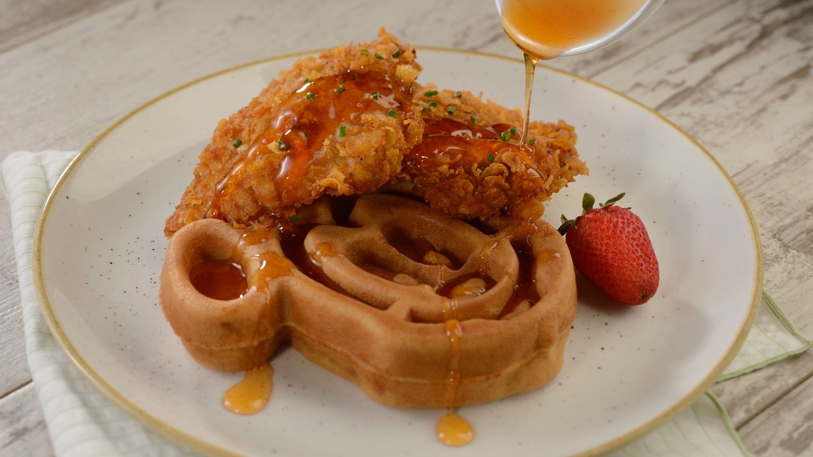 Two pieces of crispy fried chicken on a Mickey-shaped waffle with a strawberry on a white plate, as honey is poured over the top