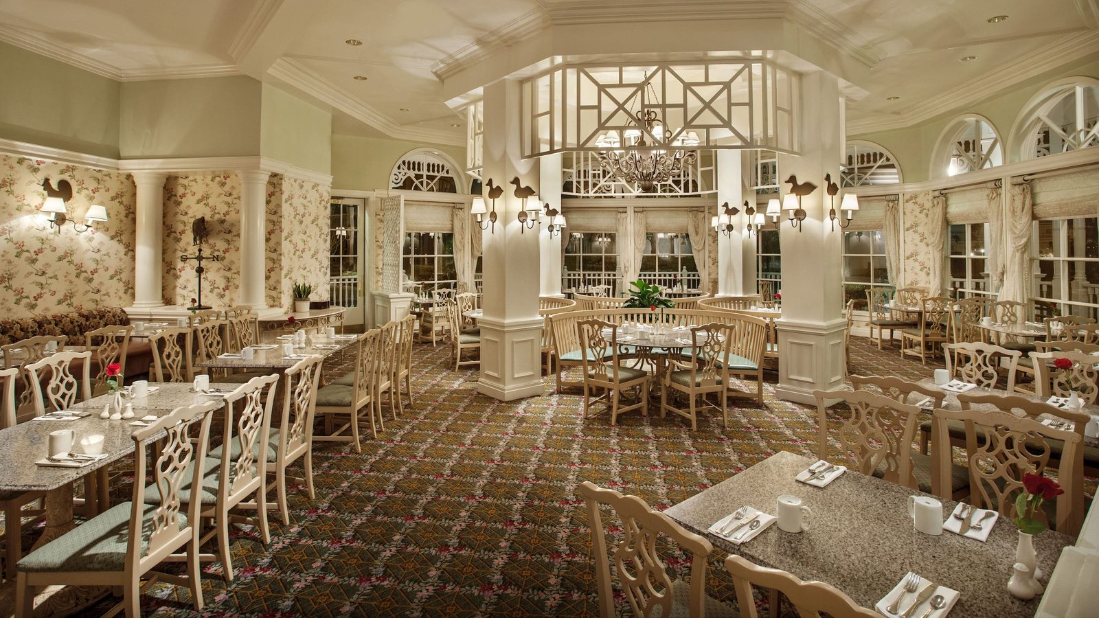 A green and cream dining room with floral wallpaper, bird-shaped wall sconces, a weathervane, white columns, granite-topped tables and a chandelier hanging inside a white lattice rotunda