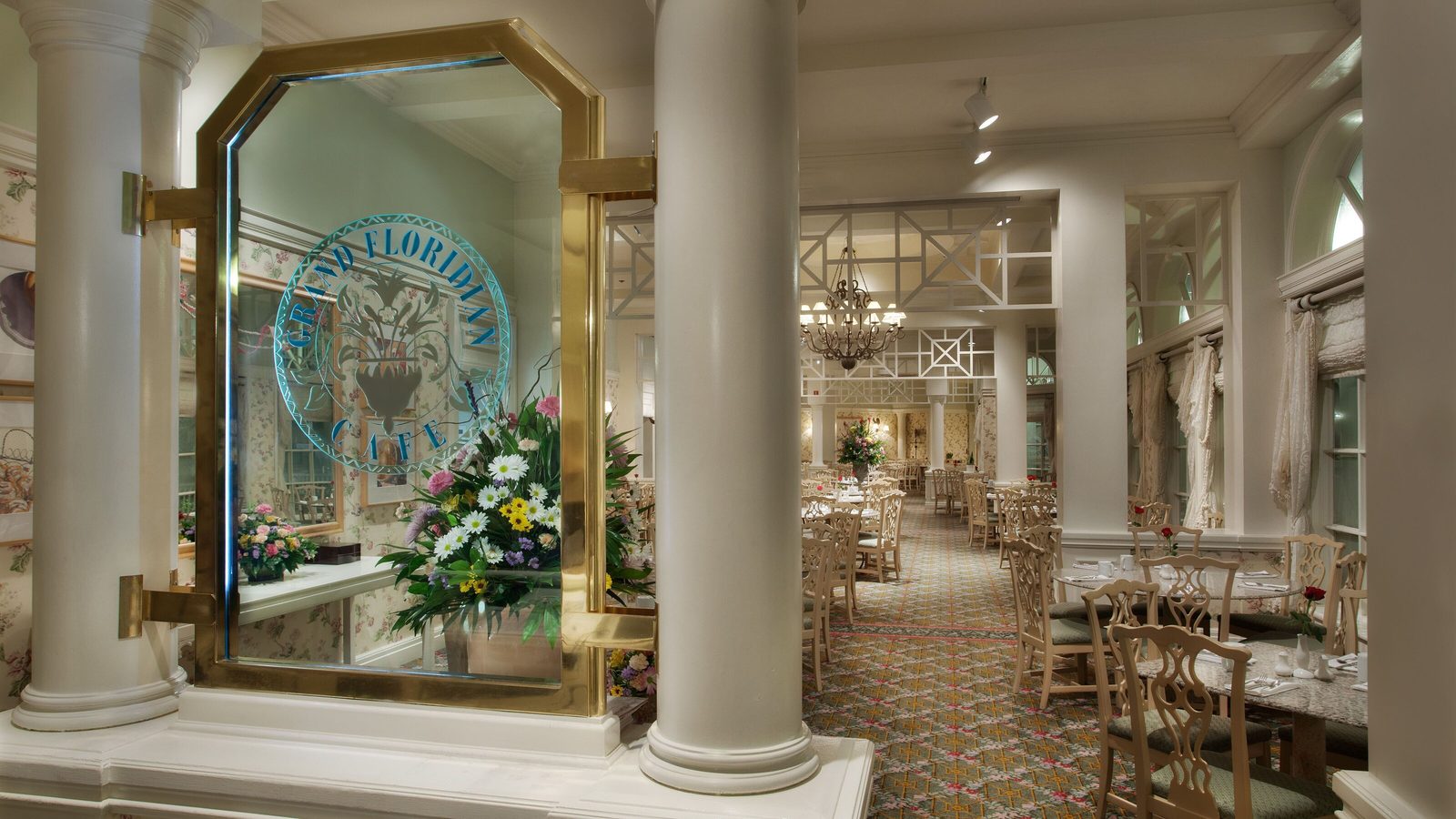 A gold-framed glass panel etched with the Grand Floridian Cafe logo beside a large flower arrangement and white columns, with the dining room and a chandelier visible behind