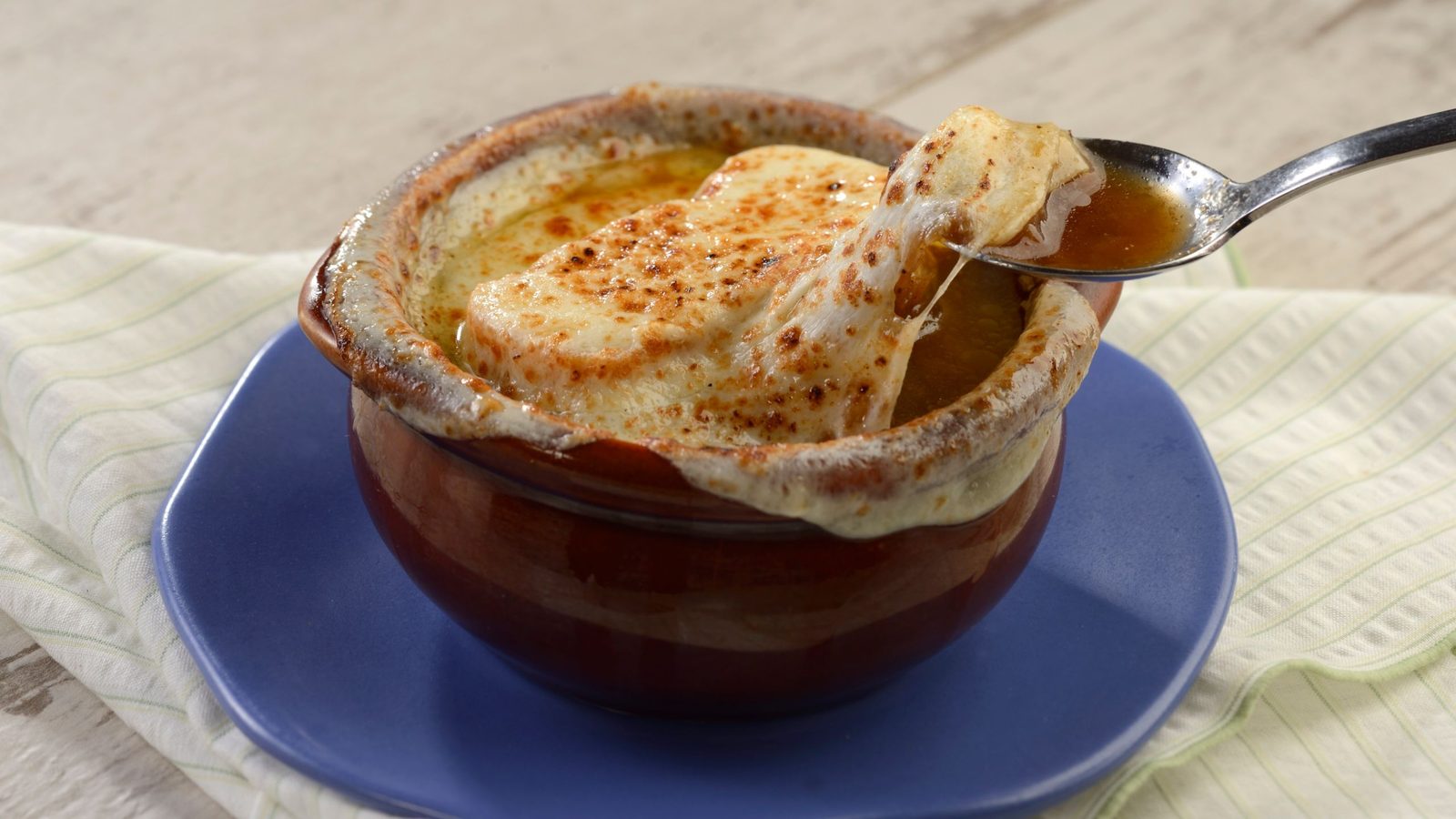 A brown crock of onion soup with a thick layer of browned melted cheese on a blue plate, with a spoon lifting a cheesy bite