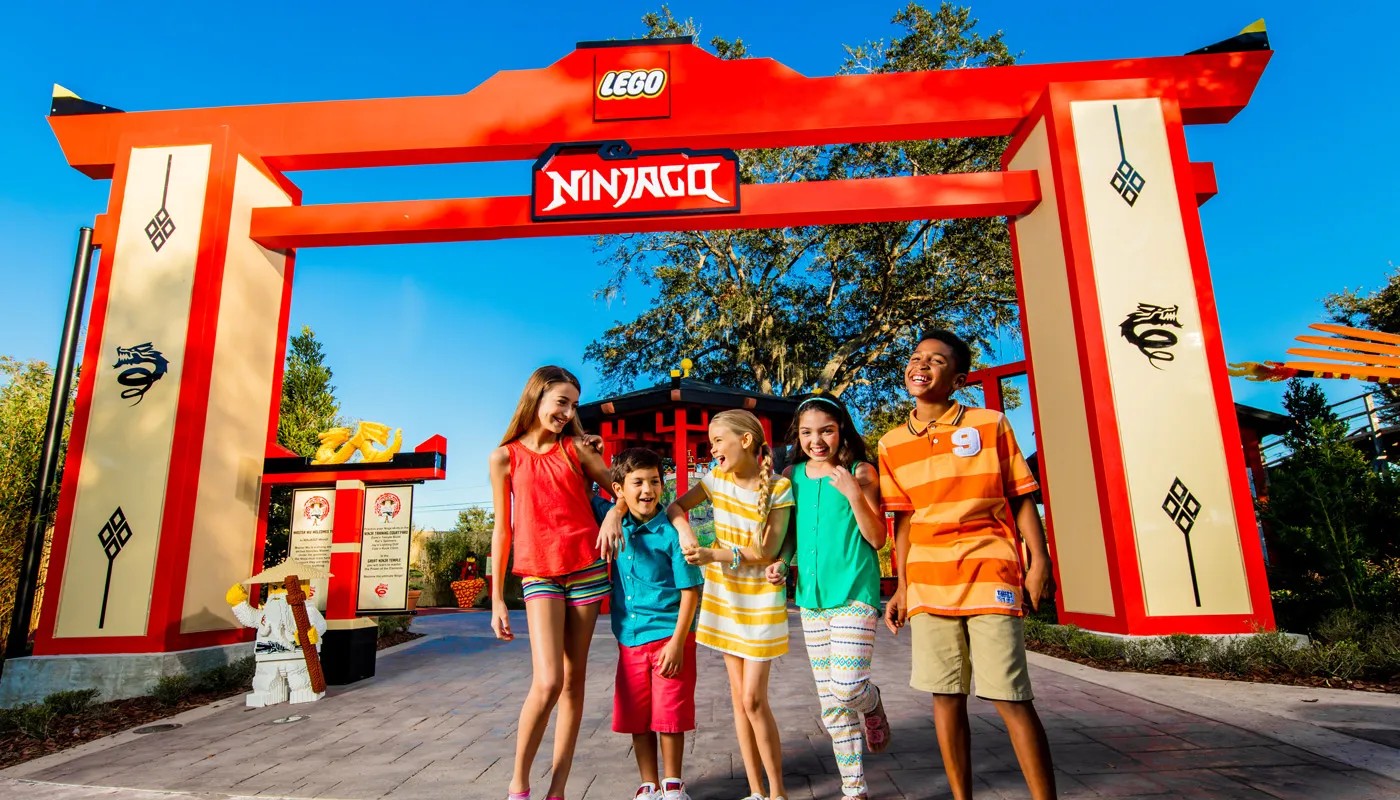 Five smiling kids walk under a large red and cream LEGO NINJAGO gateway arch, with a LEGO Master Wu figure and signs to the left and trees and blue sky behind