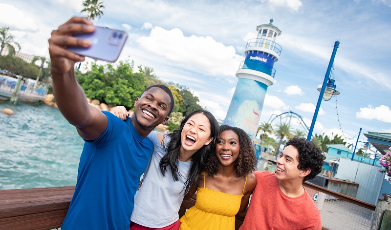 Four smiling friends take a selfie on a waterfront boardwalk, with the striped SeaWorld lighthouse and a roller coaster's blue track behind them under a blue sky