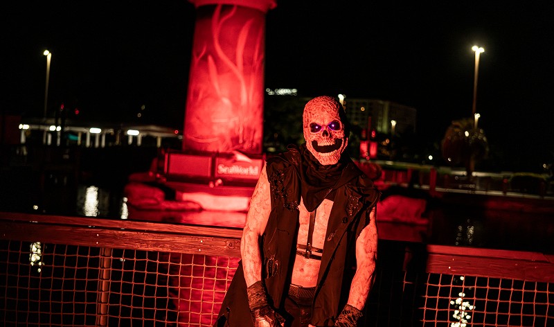 A Howl-O-Scream character in a skull mask with glowing purple eyes and a ragged black vest stands at a railing at night, with a red-lit lighthouse and a SeaWorld sign behind him