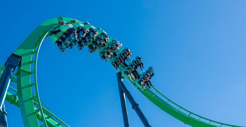 A train of riders hangs upside down through a green loop of the Kraken roller coaster against a clear blue sky