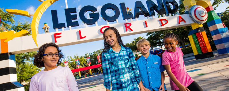 Four smiling kids in front of the colorful LEGOLAND FLORIDA entrance arch, with brick-built pillars, under a bright blue sky
