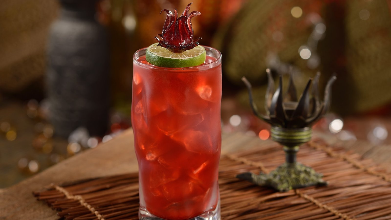 A deep red iced drink in a tall glass garnished with a lime wheel and a dark red flower, on a woven mat beside a small decorative metal stand