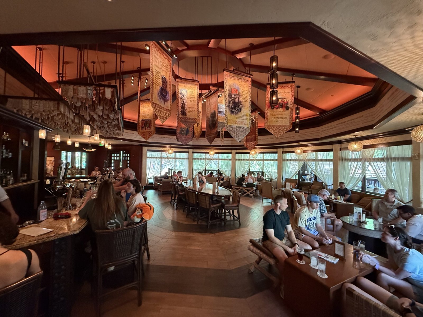 Inside Nomad Lounge: guests sit at a curved bar and low tables under an octagonal ceiling hung with colorful painted banners and lanterns, with tall curtained windows along the back wall