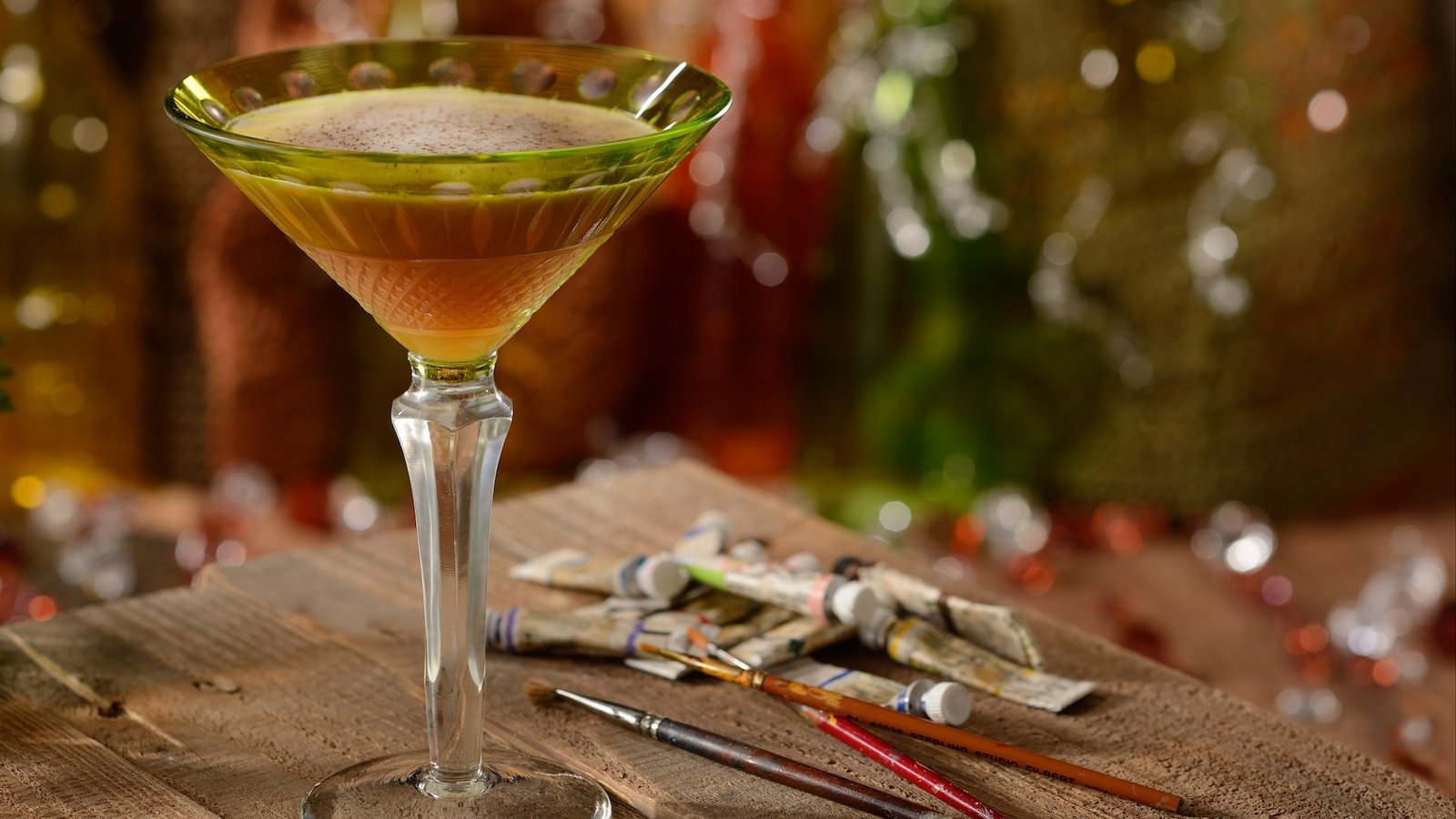 An amber cocktail with a spice-dusted foam in a green cut-glass martini glass on a wooden table, beside tubes of paint and paintbrushes