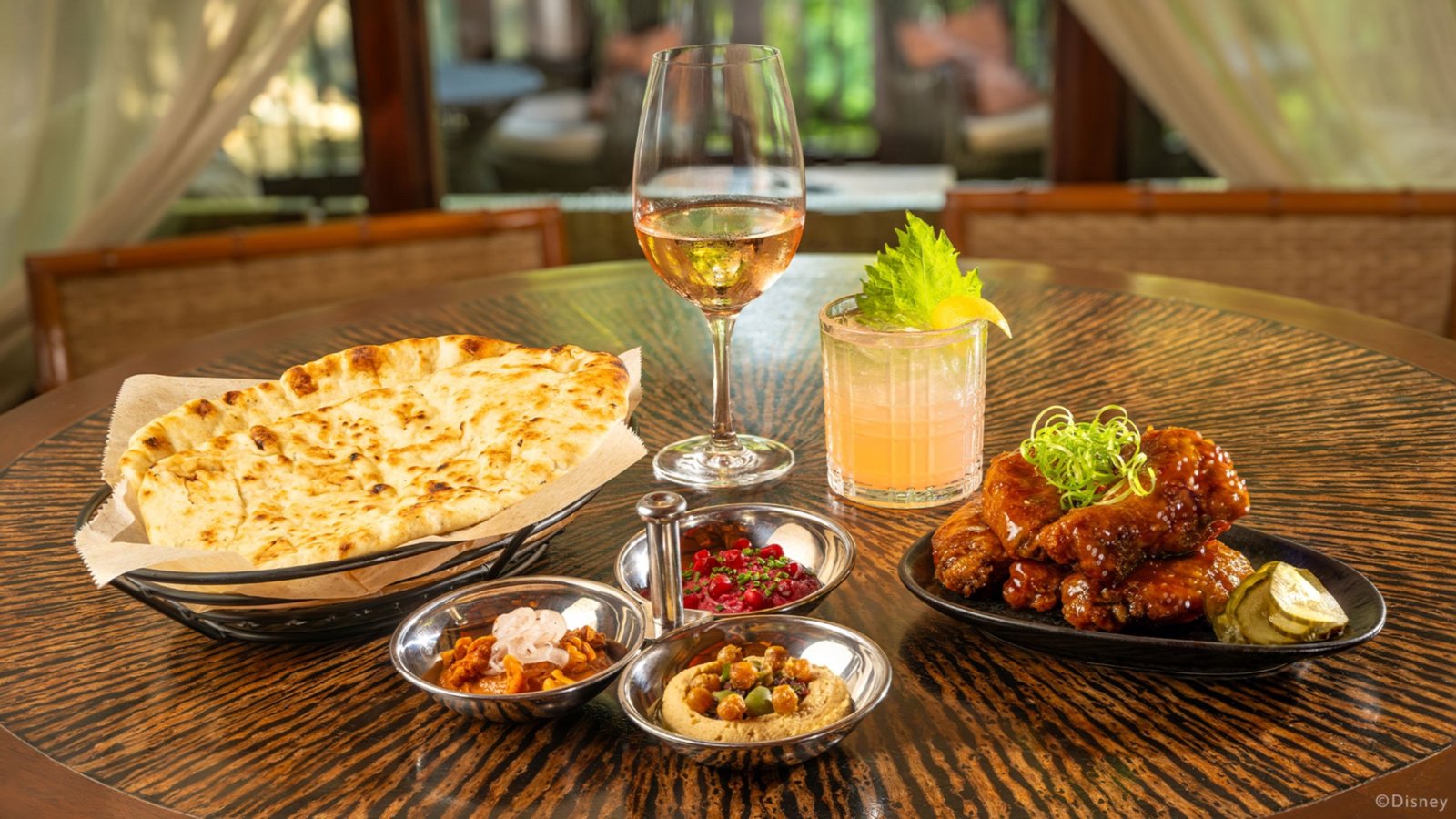 Nomad Lounge small plates on a round wooden table: a basket of warm naan, three small bowls of dips, a plate of glazed chicken wings topped with green onion, a glass of rose wine and a cocktail garnished with a leafy herb sprig and a lemon wedge