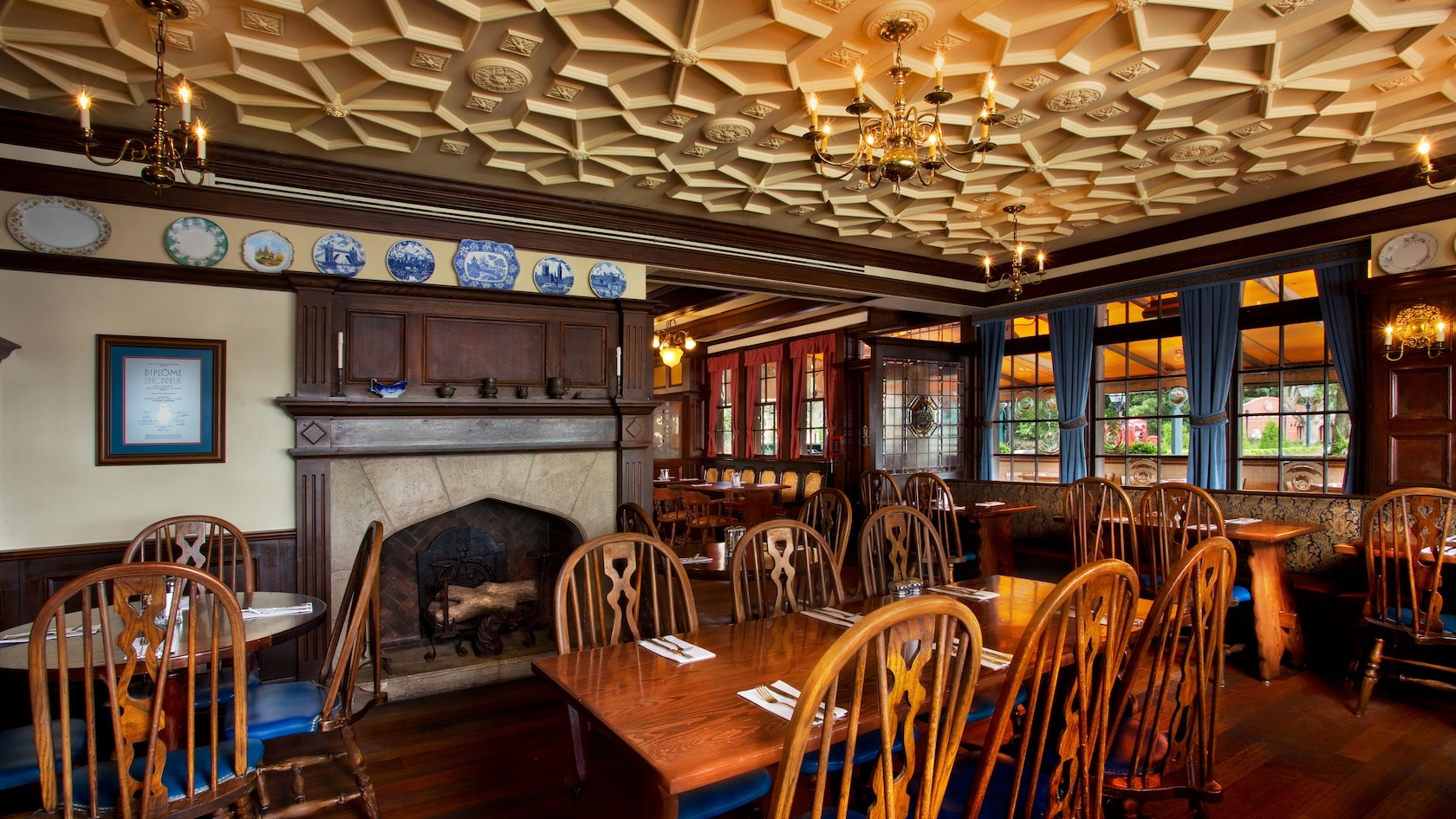 Inside the Rose & Crown Dining Room: Windsor-back chairs and wooden tables, a stone fireplace with a dark wood mantel, decorative plates (many blue and white) on a rail above, an ornate geometric plaster ceiling with brass chandeliers, and blue-curtained windows at right