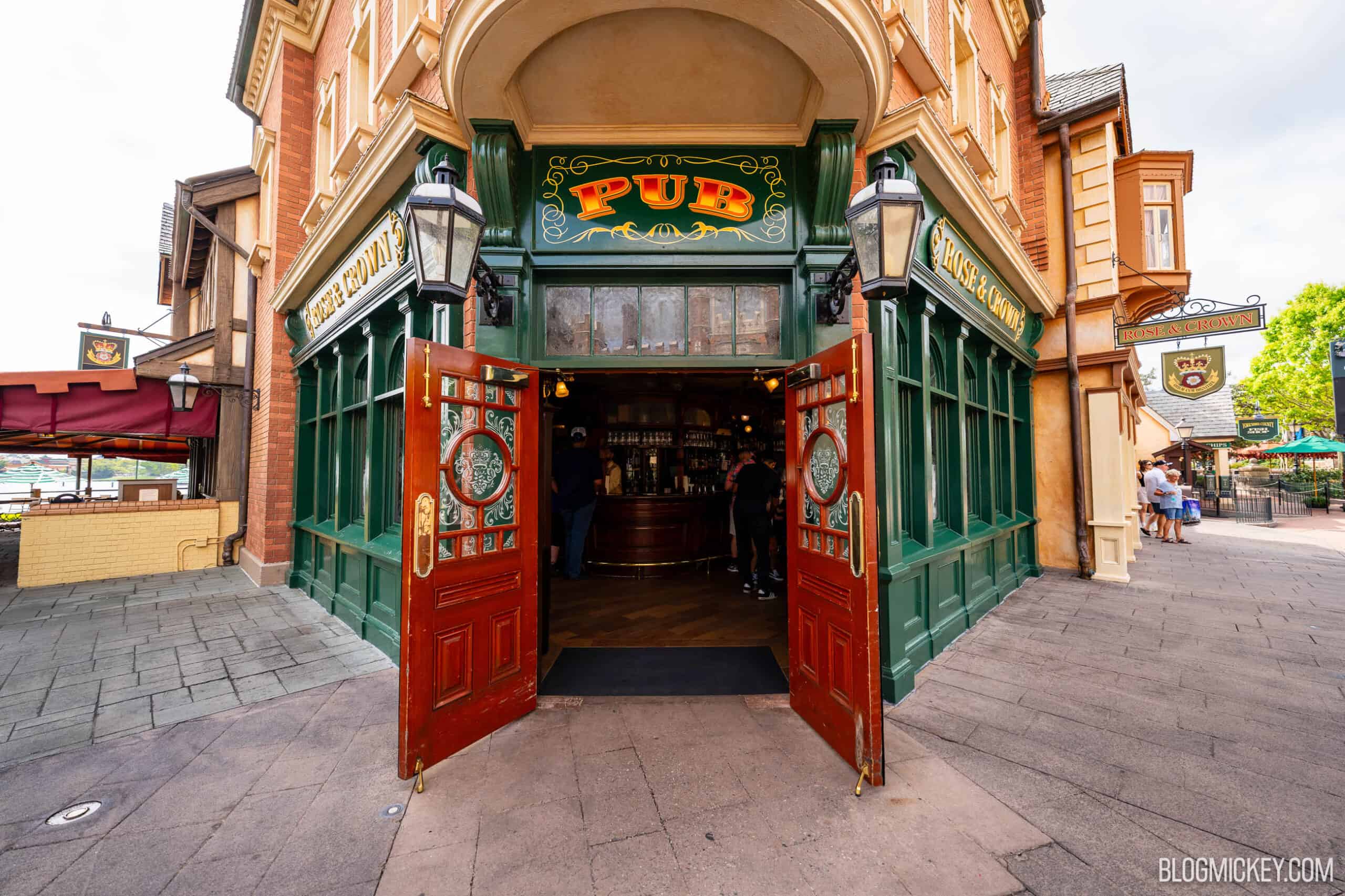 The corner entrance of the Rose & Crown Pub at EPCOT: green wooden storefront, a red and gold PUB sign above open red doors, carriage lamps either side, and guests standing at the bar inside