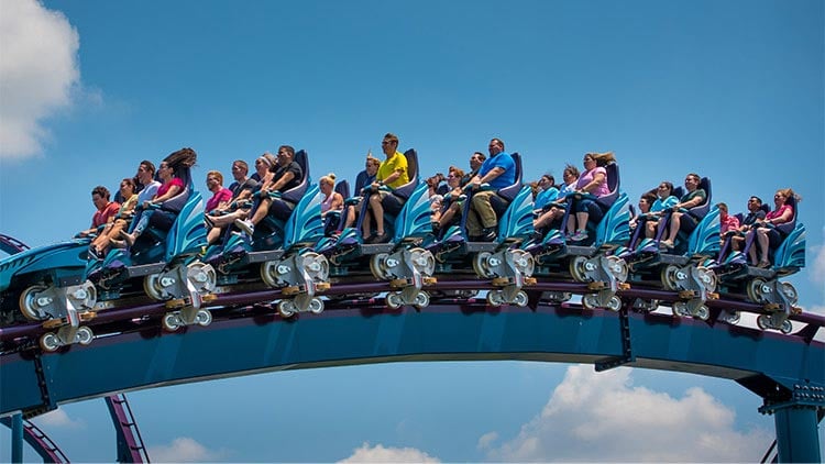 A full Mako train crests a hill against a blue sky, riders seated in open rows with lap-bar restraints, their hair blowing back