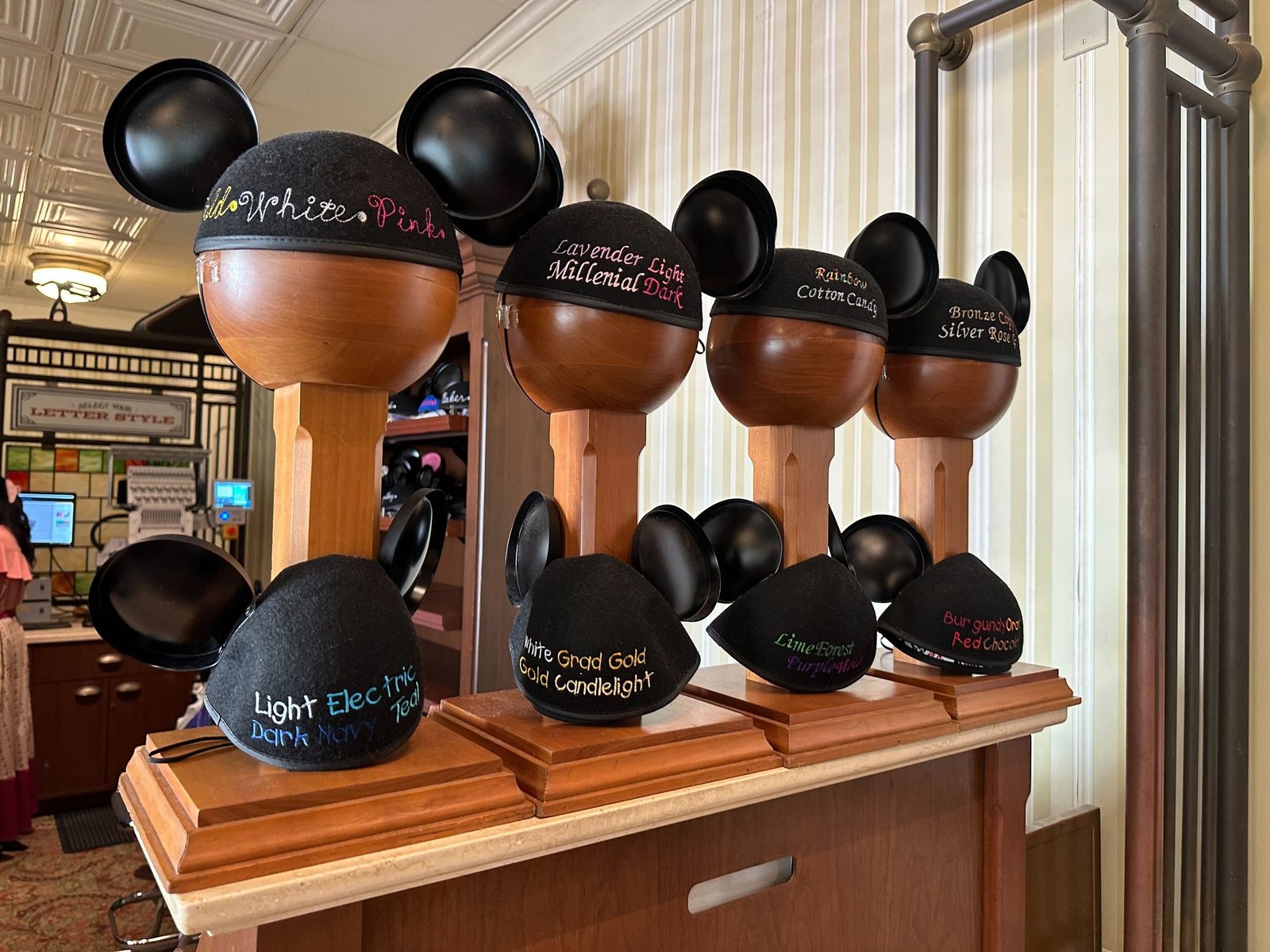 Sample black Mickey ear hats on wooden display stands, each embroidered with the names of thread colors in those colors, including Gold, White and Pink, Rainbow, Cotton Candy, Bronze, Silver, Lavender, Lime, Forest, Teal and Navy