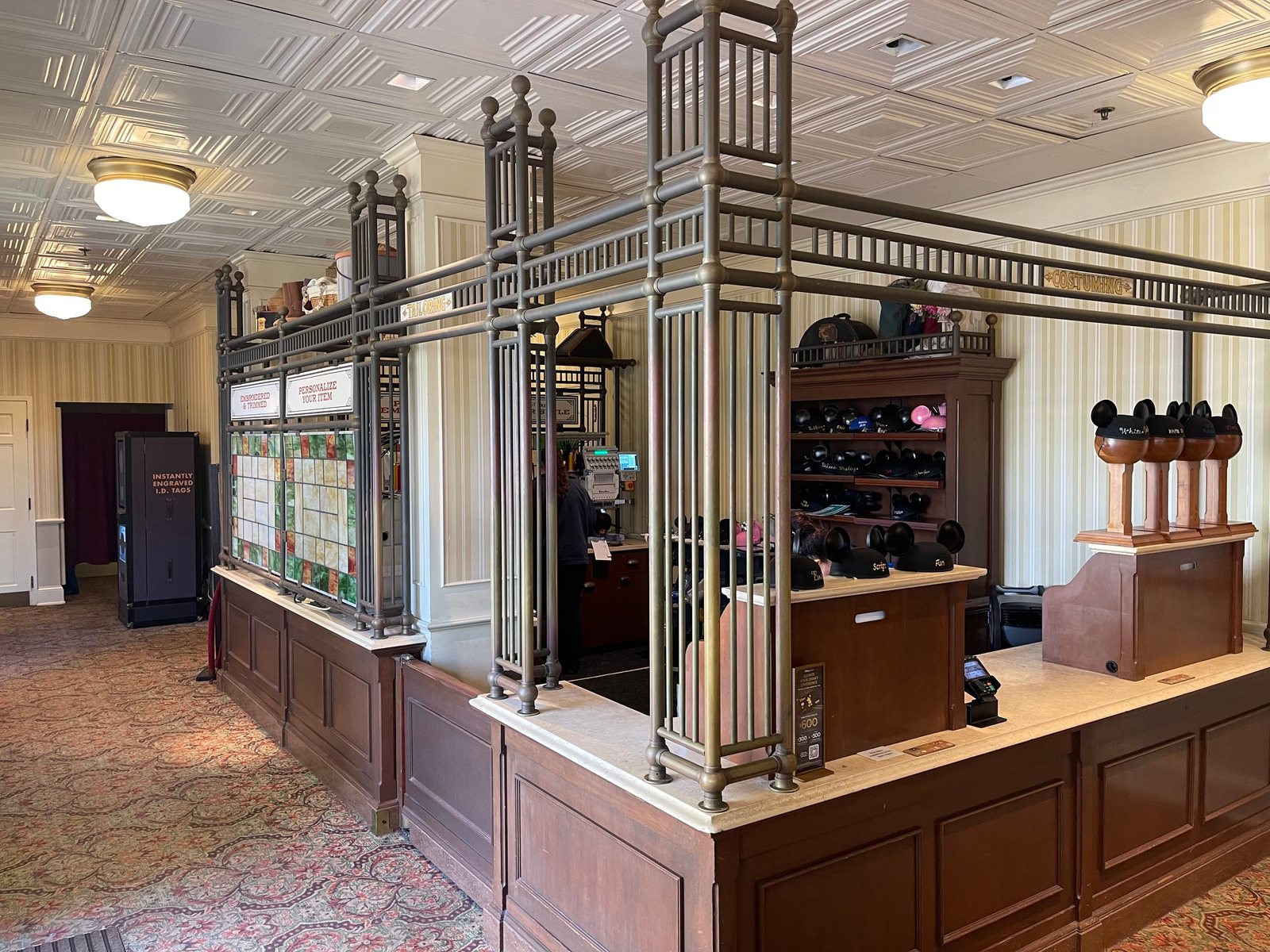 The embroidery counter inside Curtain Call Collectibles, with a bronze-colored metal frame, a Personalize Your Item sign, a wall of Mickey ear hats behind the counter and ear hats on wooden stands, under a pressed-tin ceiling